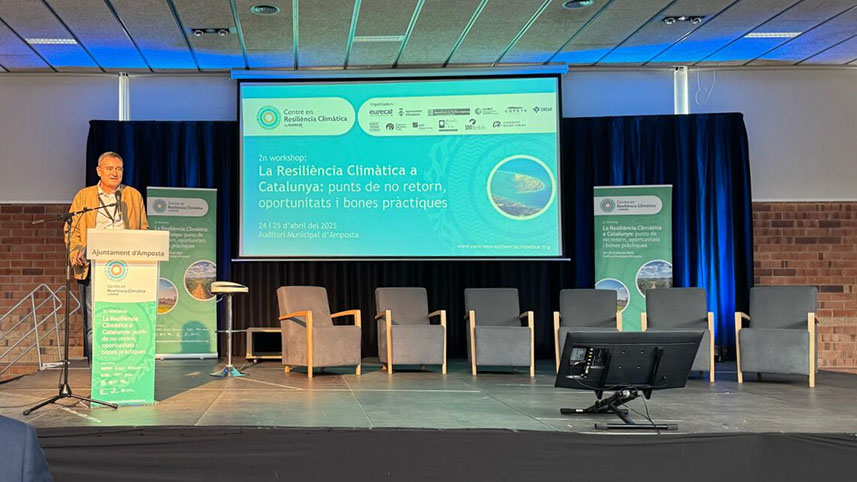 A speaker speaks from a lectern on a stage with a large screen behind him showing the title of the workshop on climate resilience in Catalonia, with several chairs set up for a round table.