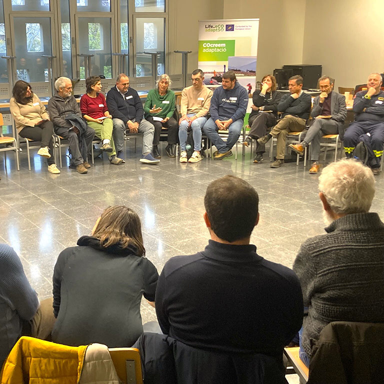 Group of people sitting in a circle in a room, participating in a dialogue space of the Life eCOadapt50 project, with an information poster about the project in the background.