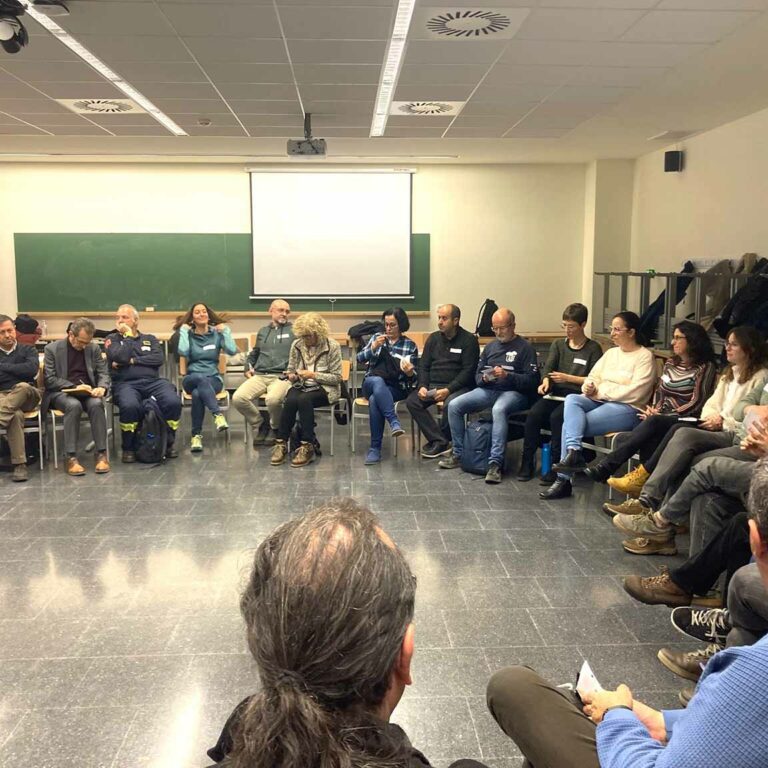 People sitting in a circle in a classroom with a green board and projector, participating in a plenary session of dialogue and sharing.
