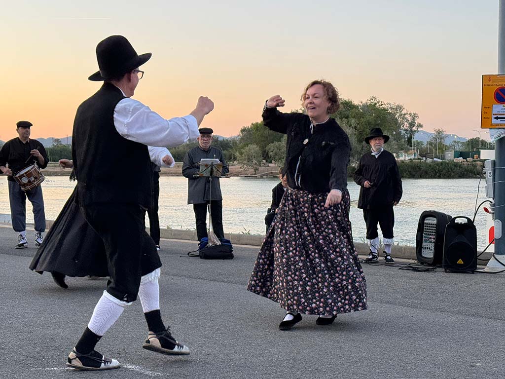 Couple dancing dressed in traditional Catalan clothing while a group of musicians play folkloric instruments next to the Ebro River, during sunset.
