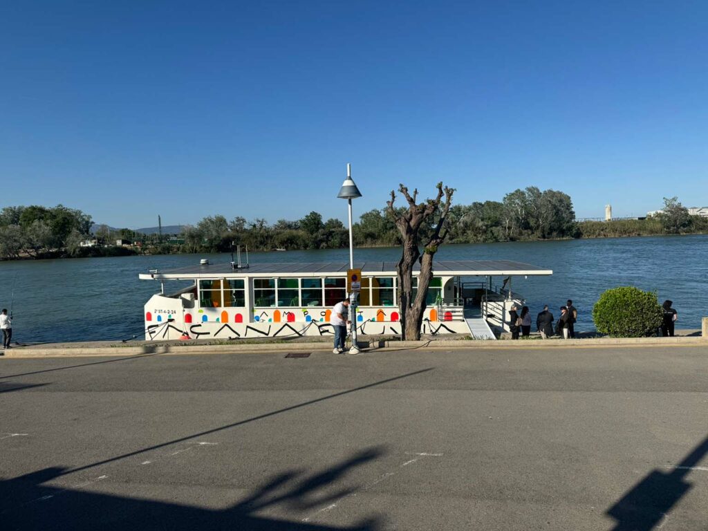 Colorfully decorated river boat docked at the Ebro River pier in Amposta, with several people nearby and a clear sky in the background.