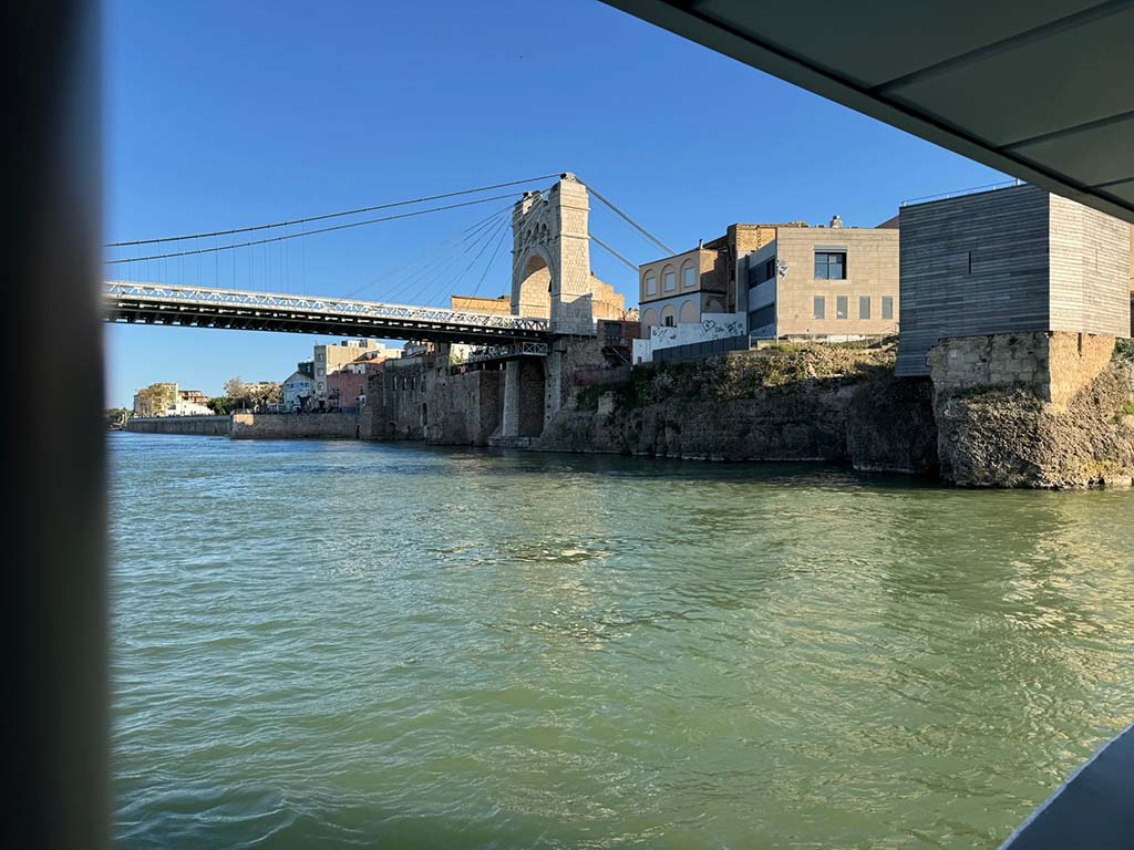 View from the Ebro River of the Amposta suspension bridge with historic and modern buildings in the background, under a clear and sunny sky.
